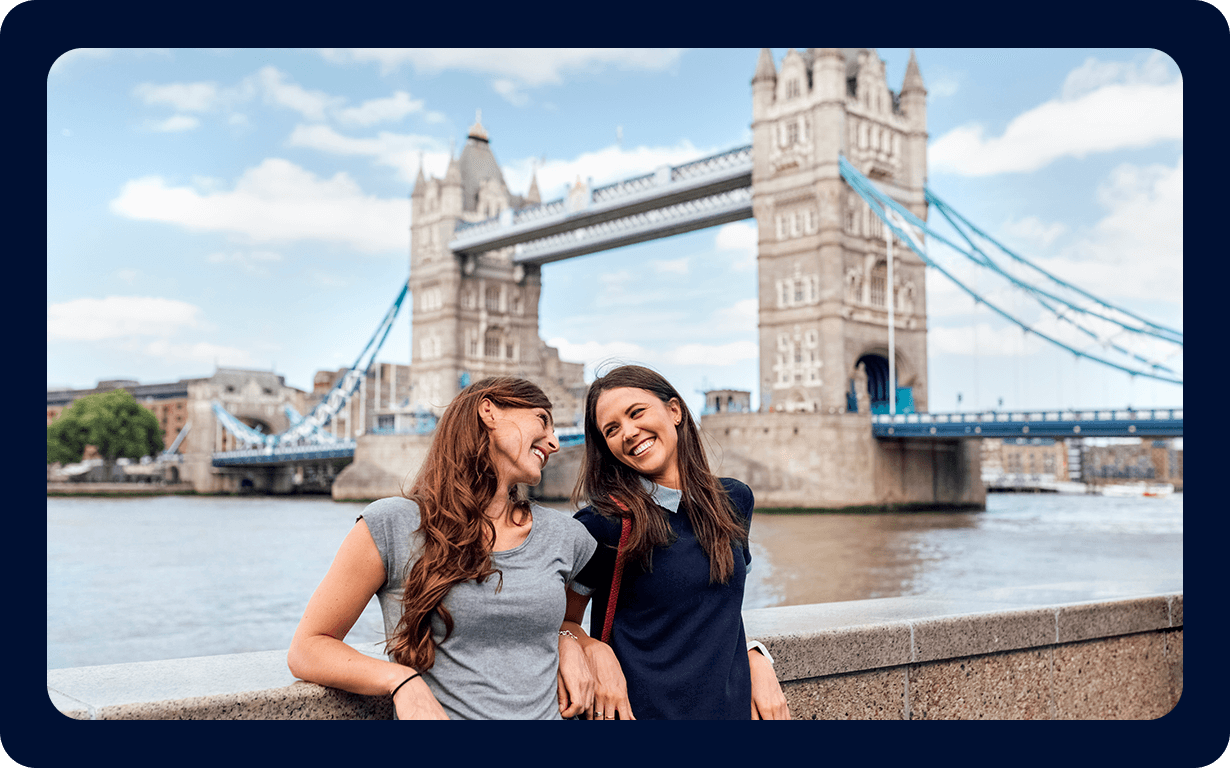 Two smiling women stand in front of the Tower Bridge in London, leaning on a stone railing as they enjoy the view and each other's company. The sky is partly cloudy, and the iconic bridge forms a picturesque background—a perfect moment for celebrating their decision about moving to the UK.