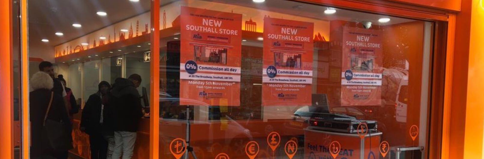 Bright orange storefront with large windows, advertising "New Southall Store" and "0% Commission All Day" in multiple posters. Several people are inside the store, lined up at the counter. Currency symbols are displayed on the exterior window frame.