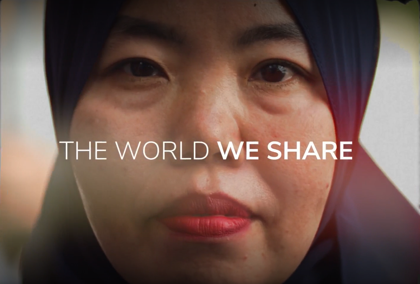 A close-up of a woman wearing a dark-colored hijab and red lipstick. Text overlaying the image reads, "THE WORLD WE SHARE." The background is slightly blurred, drawing focus to the woman's face and the text.