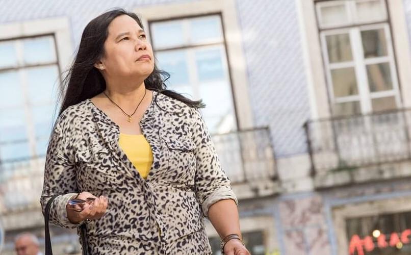 A mother from the Philippines with long dark hair, wearing a leopard print blouse over a yellow top, walks outside holding a cell phone. She looks upwards with a serious expression. Behind her, there are buildings with large windows and balconies.
