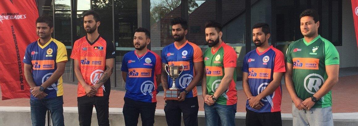 A group of seven men in colorful sports jerseys stands in a line outdoors. The man in the center holds a large trophy. They appear to be part of a sports team, possibly cricket, given the logos and context. A red flag and a building are visible in the background.