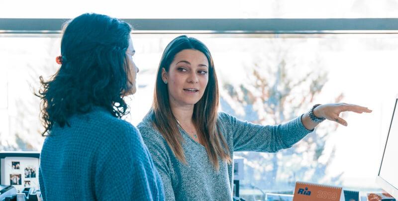 Two women in technology are engaged in a discussion in an office setting. One woman is gesturing towards a computer screen, explaining something to the other. Both are dressed in casual attire, and there is a window with a bright, blurred outdoor view in the background.