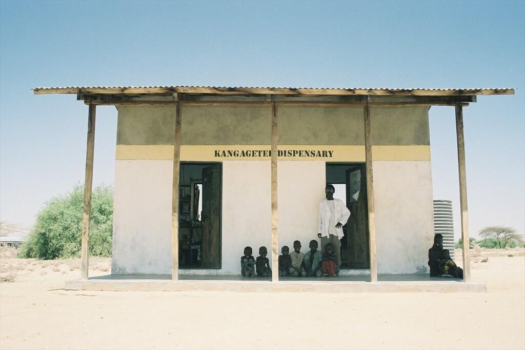 A small rural building with a sign reading "Kangagetei Dispensary" has several children sitting outside on the porch. An adult in a white coat stands in the doorway, potentially discussing remittances and health coverage. The surrounding area is dry and sparsely vegetated, under a clear blue sky.