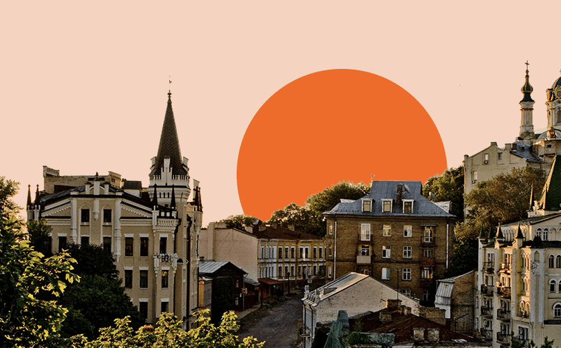 A picturesque view of a historic European cityscape at sunset. The scene features a mix of old buildings with spires and classic architecture, with a large orange sun setting in the background, casting a warm glow over the landscape. Trees are interspersed among the buildings.