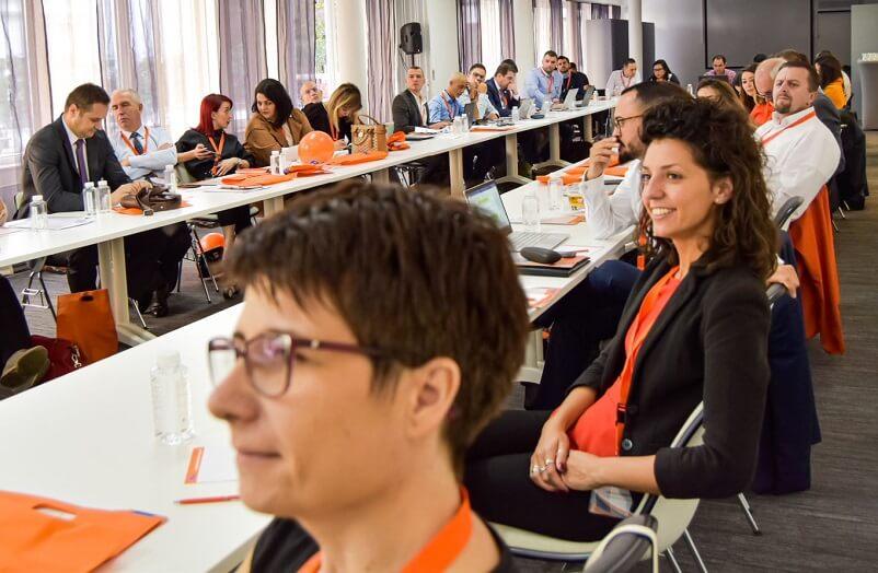 A room filled with people seated around a large, U-shaped conference table, engaged in discussions. Most attendees have laptops, notepads, and orange lanyards. The room is well-lit with large windows on one side. A woman in the foreground smiles, facing the camera.