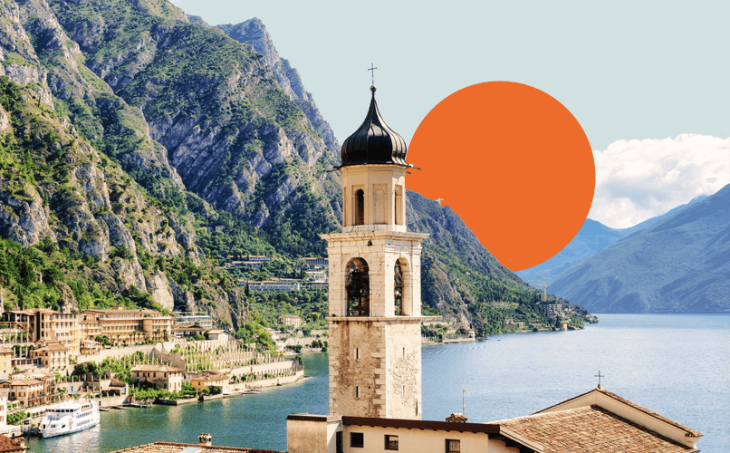 A picturesque view of a lakeside town nestled among steep, green mountains. A tall bell tower stands prominently in the foreground, with the serene lake and a vibrant, orange sun setting behind the mountains in the background, whispers stories of migration and remittances in Italy's past and present.