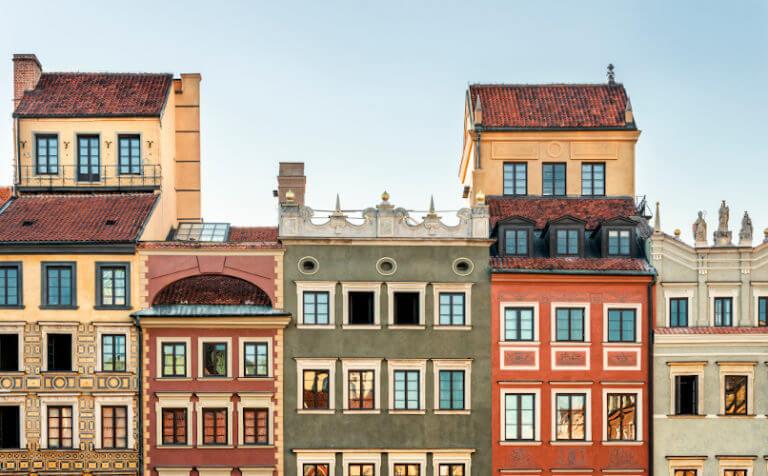Row of colorful historic buildings with intricate architectural details and gable roofs, set against a clear blue sky. The facades feature various window designs and decorations, showcasing a blend of colors including beige, green, red, and yellow—reminiscent of charming streets in Poland.