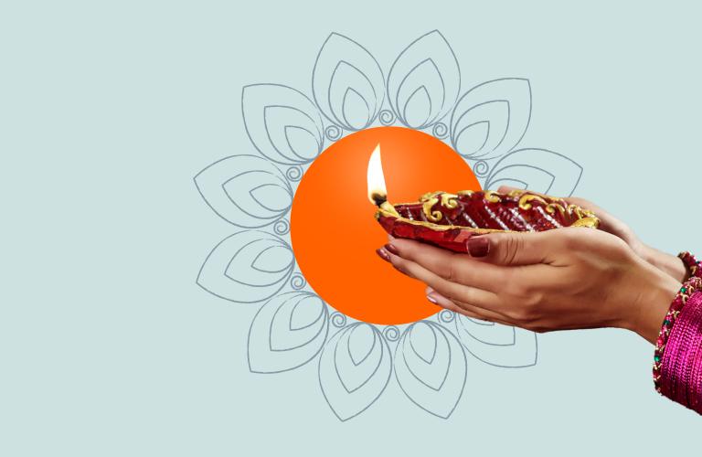 A pair of hands holding a lit clay lamp with a flame. The hands are adorned with red bangles. In the background, there's an orange circle with an intricate floral pattern surrounding it, set against a light blue backdrop.