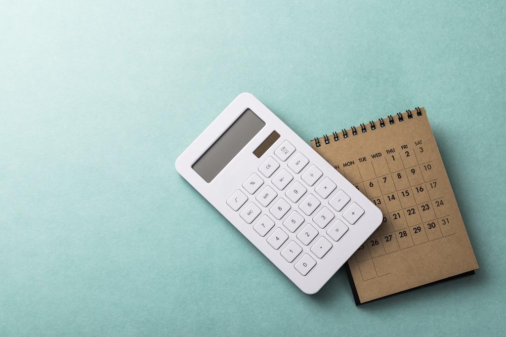Una calculadora blanca se encuentra sobre una superficie azul claro junto a un pequeño calendario marrón con espiral abierto para mostrar un mes. La calculadora está apagada y el calendario muestra las fechas en formato de cuadrícula. Los objetos están dispuestos uno al lado del otro.