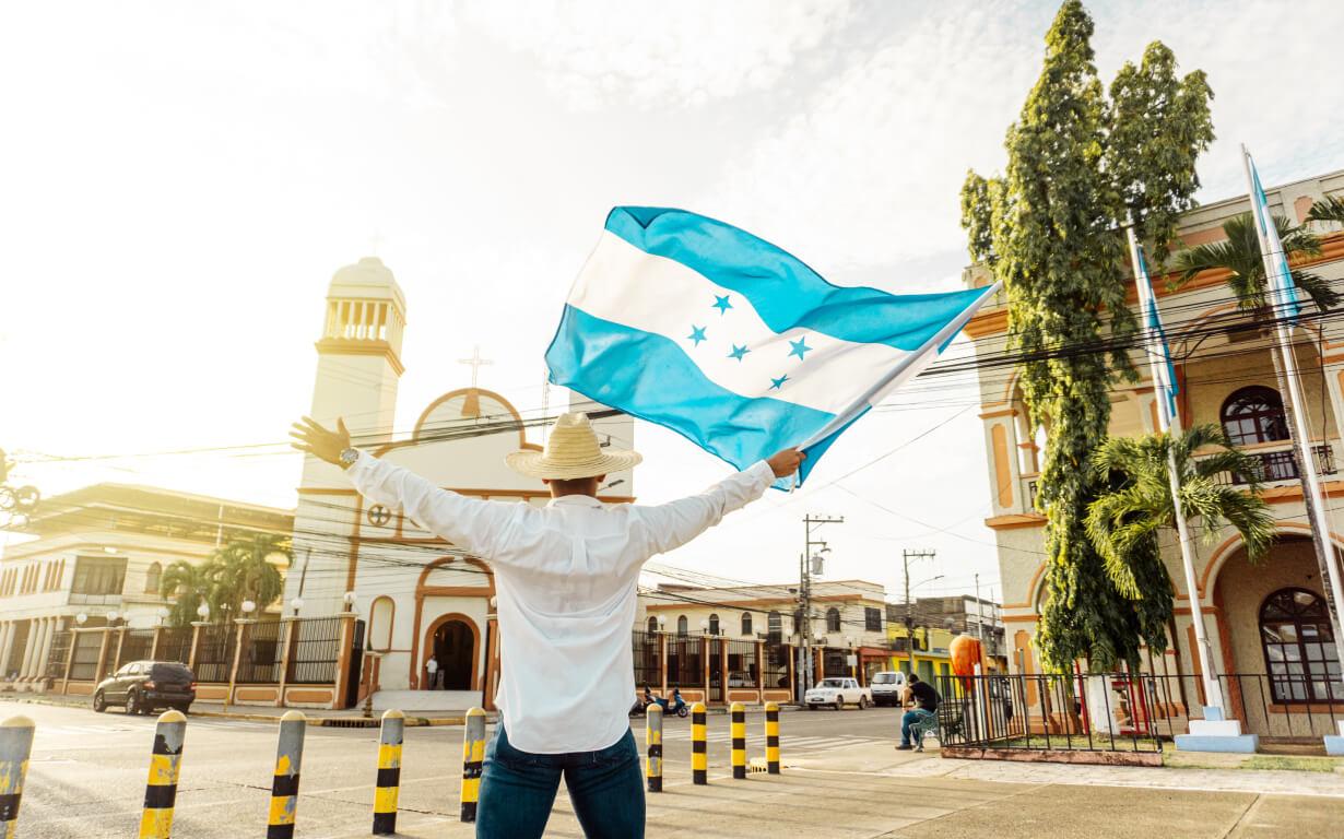Une personne portant un chapeau et une chemise blanche agite un drapeau à rayures bleues et blanches et cinq étoiles bleues devant une église et un bâtiment dans une rue bordée de bornes jaunes et noires, commémorant le 15 septembre par une journée lumineuse et ensoleillée.