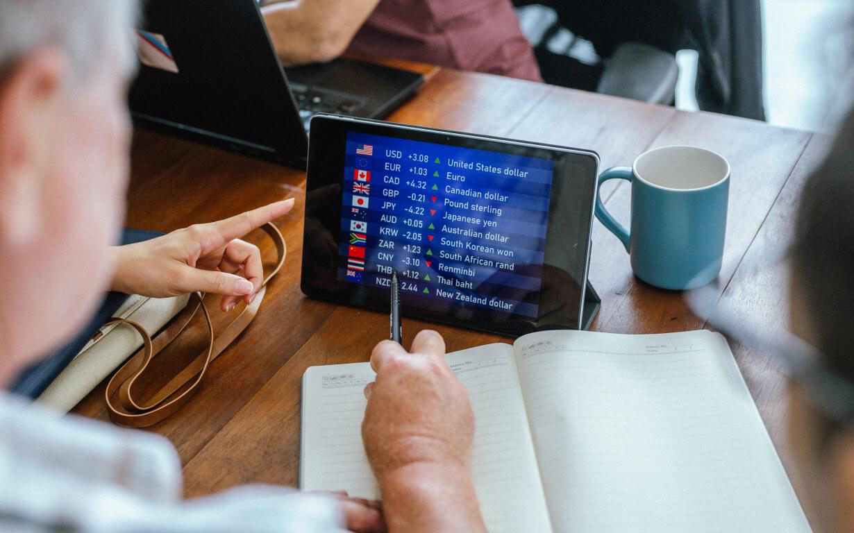Dos personas están sentadas en una mesa con cuadernos. Una de ellas señala una tableta que muestra los tipos de cambio de divisas, incluidos el dólar estadounidense, el euro y la libra esterlina. Sobre la mesa hay una taza azul.