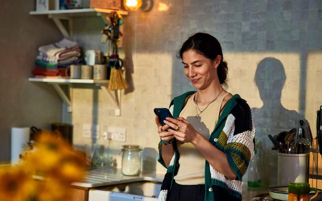 A woman in a cozy kitchen looks at her phone, smiling as she explores the safest ways to send money. She wears a knitted sweater over a beige top. Sunlight casts shadows on the tiled wall behind her, with shelves of cups and folded towels in the background and yellow flowers in the foreground.