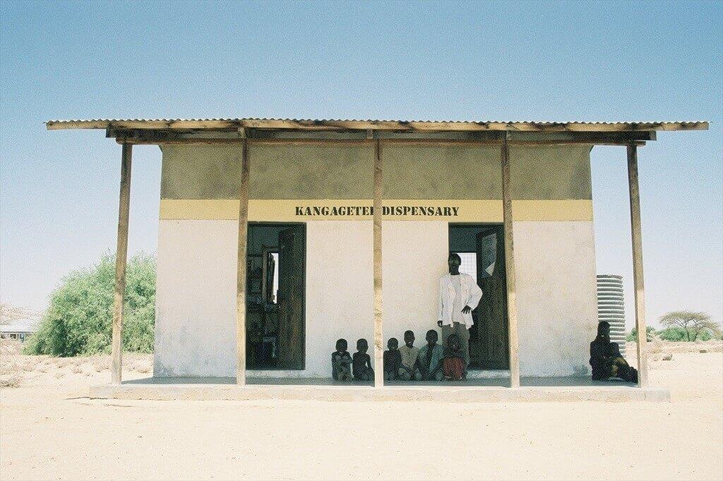 Un pequeño edificio, llamado "Dispensario Kangagetei", se alza como un rayo de esperanza. Varias personas, incluidos niños, esperan bajo el alero los servicios de salud que tanto necesitan en esta zona seca y arenosa. Gracias a las remesas y a la mejora de la cobertura sanitaria, muchos ahora tienen acceso a atención médica esencial bajo un cielo despejado.