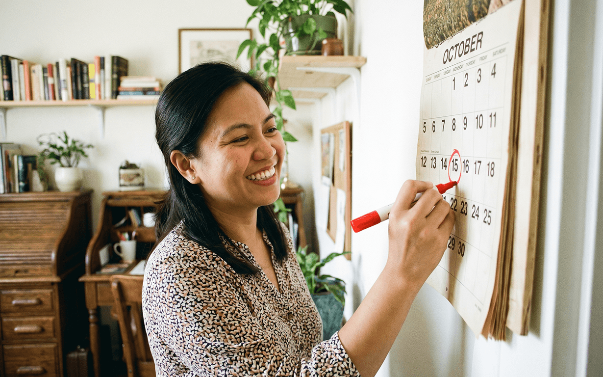 A woman smiles while circling October 16th on a wall calendar with a red marker, likely marking her money sending routine, in a cozy, plant-filled room with bookshelves and a wooden desk in the background.